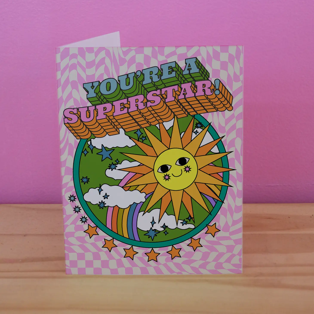 A greeting card that reads "You're A Superstar", with a wavy, pink and white checkered background and illustration of a smiling sun with star-rougued cheeks.  Behind the sun is a green sky with clouds, stars, and a rainbow.